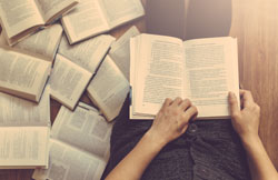 Visão de cima de uma pessoa lendo um livro e com diversos outros livros abertos ao seu lado.