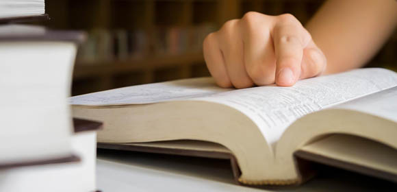 Mão repousa sobre livro aberto em uma mesa, enquanto pessoa lê atentamente uma página